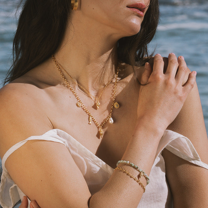 Gold-toned chain necklace with opal conch shell pendant worn layered at various lengths on a person near water.
