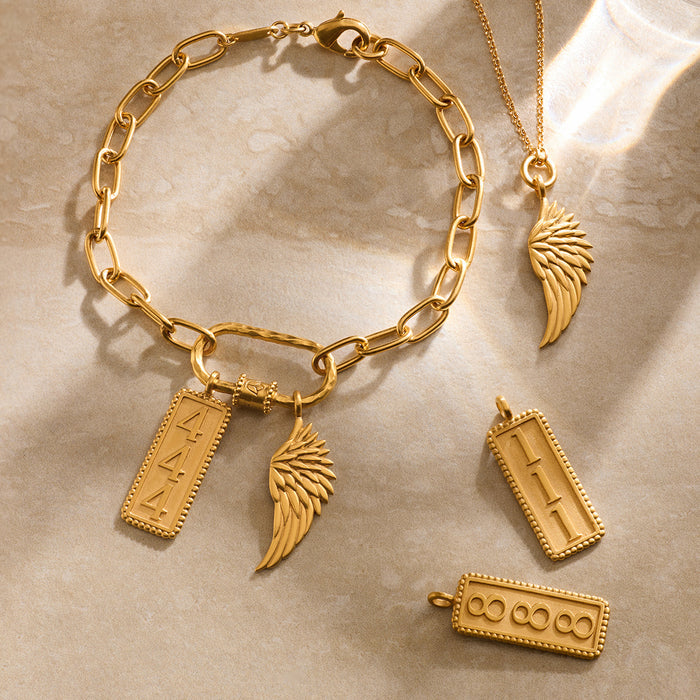 Gold-toned charm collection featuring angel wings, rectangular pendants with etched details, and a chain bracelet on neutral