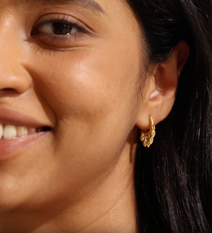 A close-up of a woman smiling, displaying the Scatter Light Huggie Earrings. These earrings embody spiritual essence and serve as a talisman for clarity and awareness, aligning with the themes of zodiac energy, meditation, and meaningful intentions in one’s spiritual journey.