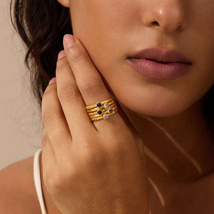 Gold stacked rings with dark and light gemstones worn on finger.