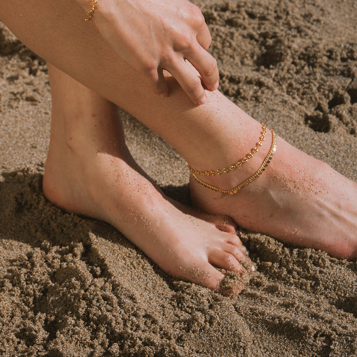 Gold chain anklets with coin charms worn on foot in sand.