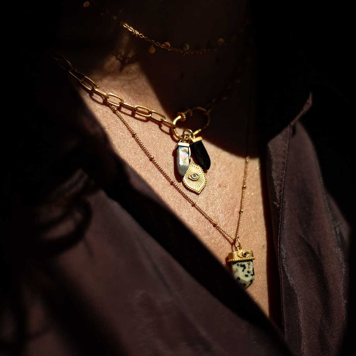 Gold charm pendant with iridescent stone and eye motif on layered chain necklace worn on person.