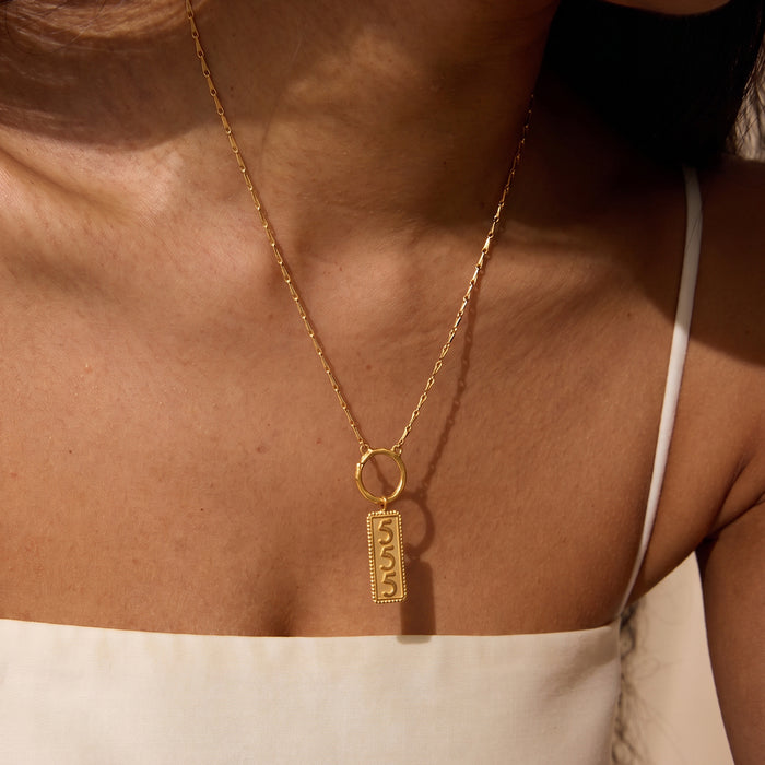 Gold-toned necklace with rectangular pendant engraved with the number 555, worn on bare chest.