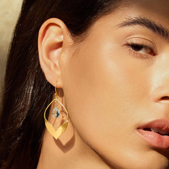 Close-up of a model wearing Transformed by Compassion Lotus Blue Topaz Earrings, featuring an elegant drop design in 18kt gold plating with blue topaz accents, symbolizing new beginnings and spiritual growth. Ideal for meditation and yoga enthusiasts, these earrings serve as a meaningful talisman for manifestation and protection.