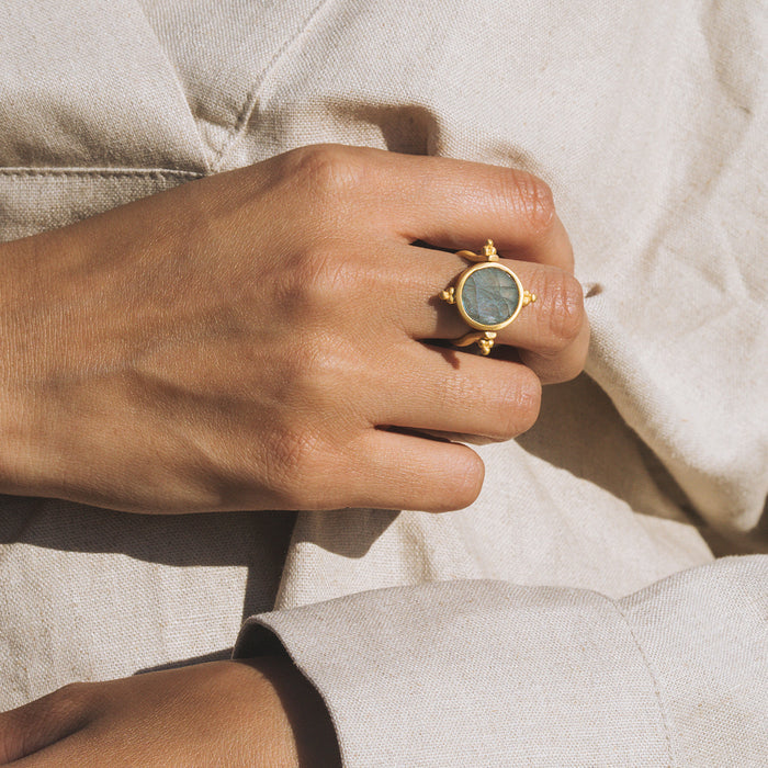 Gold-tone spinner ring with gray labradorite stone center and beaded details, worn on hand against cream fabric.