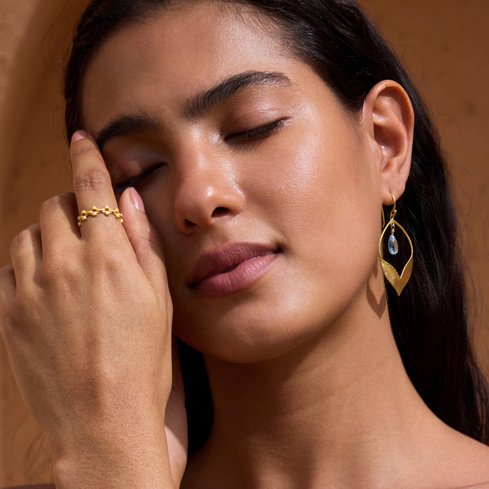 Gold textured band ring worn on ring finger of model's hand against neutral tan background.